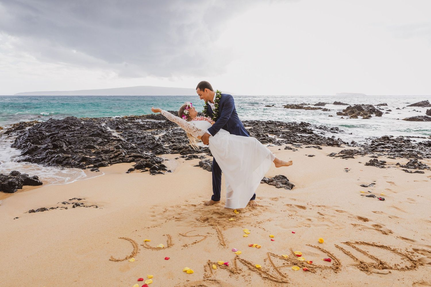Beach proposal scene with 'Marry Me' written in sand near rocky coastline.