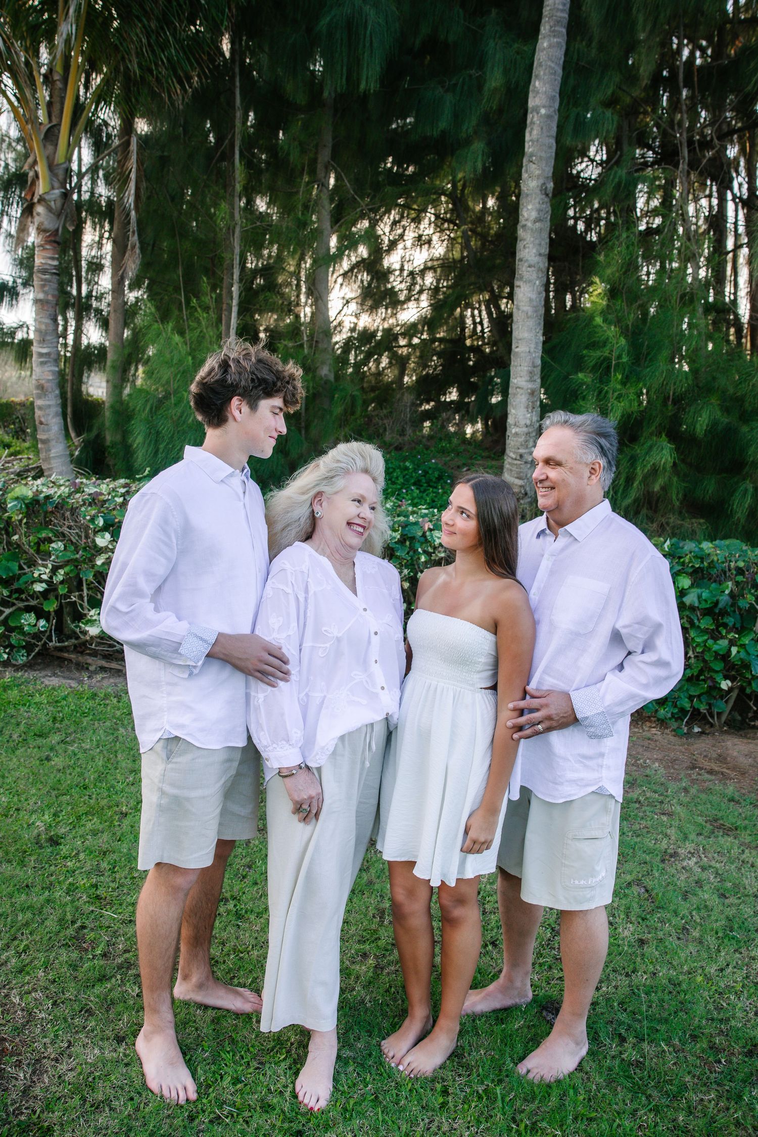 Family group wearing all white outfits posing casually on grass under palm trees.