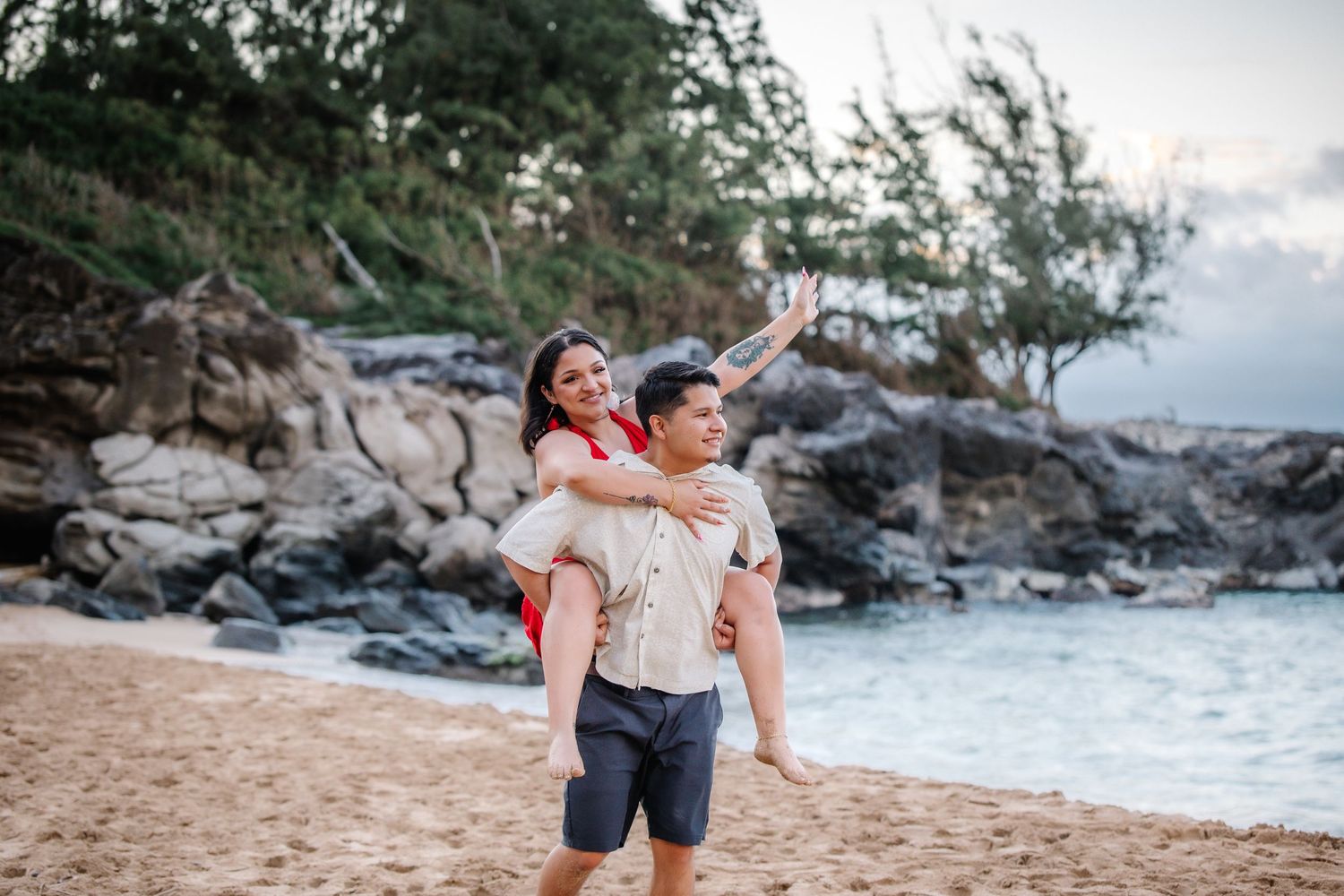 Couple enjoying a piggyback ride on a sandy beach with rocky coastline and lush trees in the background.