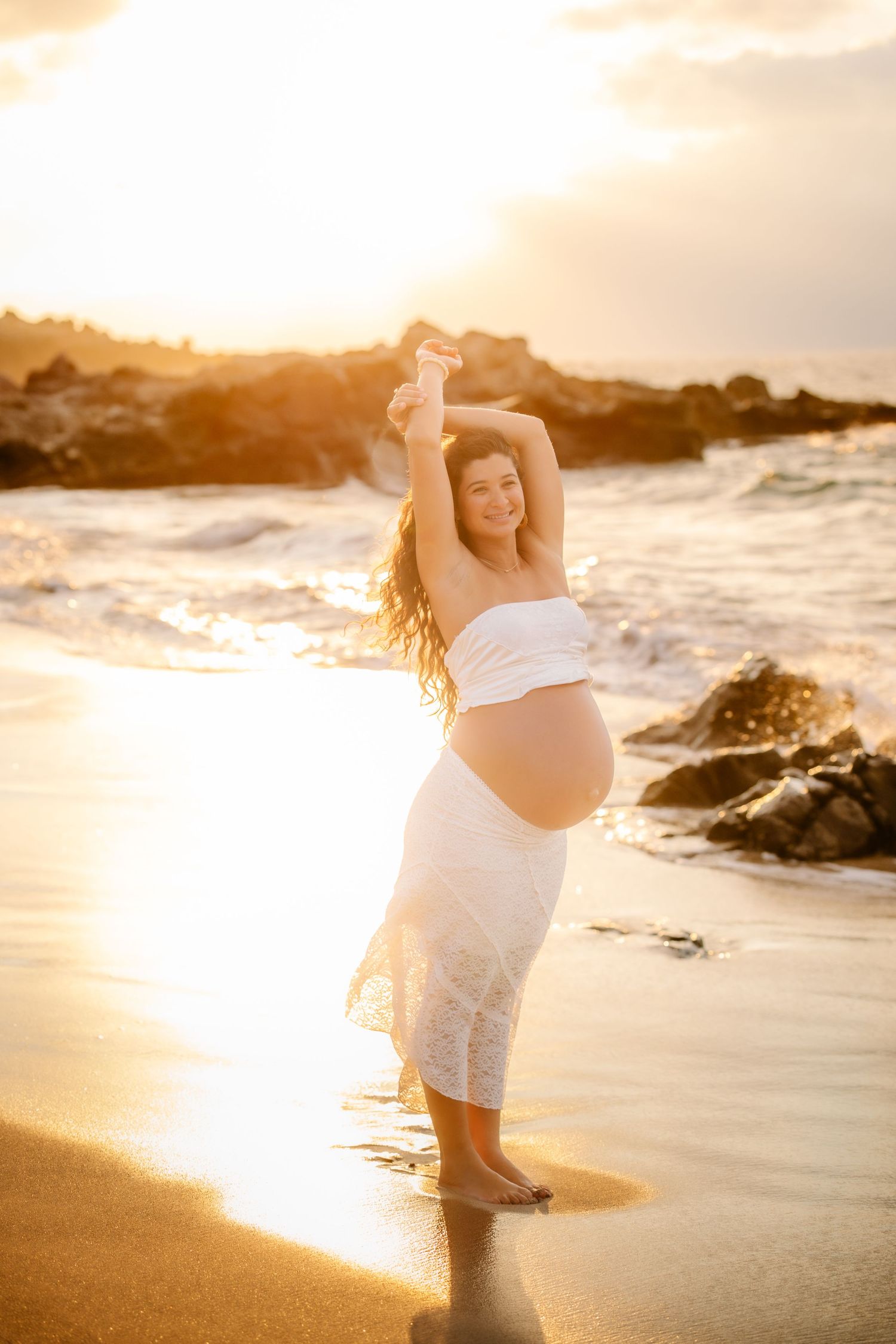 Pregnant woman in white dress poses gracefully on a golden sunset beach with waves and rocks behind her.