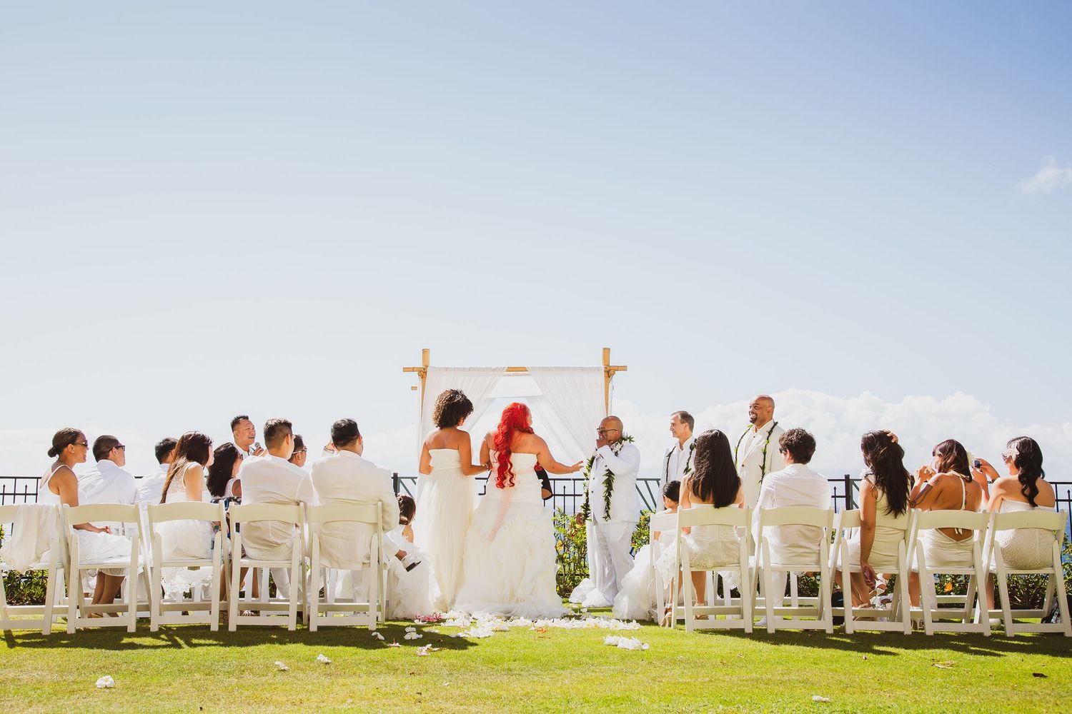 Outdoor wedding ceremony on green lawn with white chairs, wooden arch, bride and groom, and guests under clear blue sky.