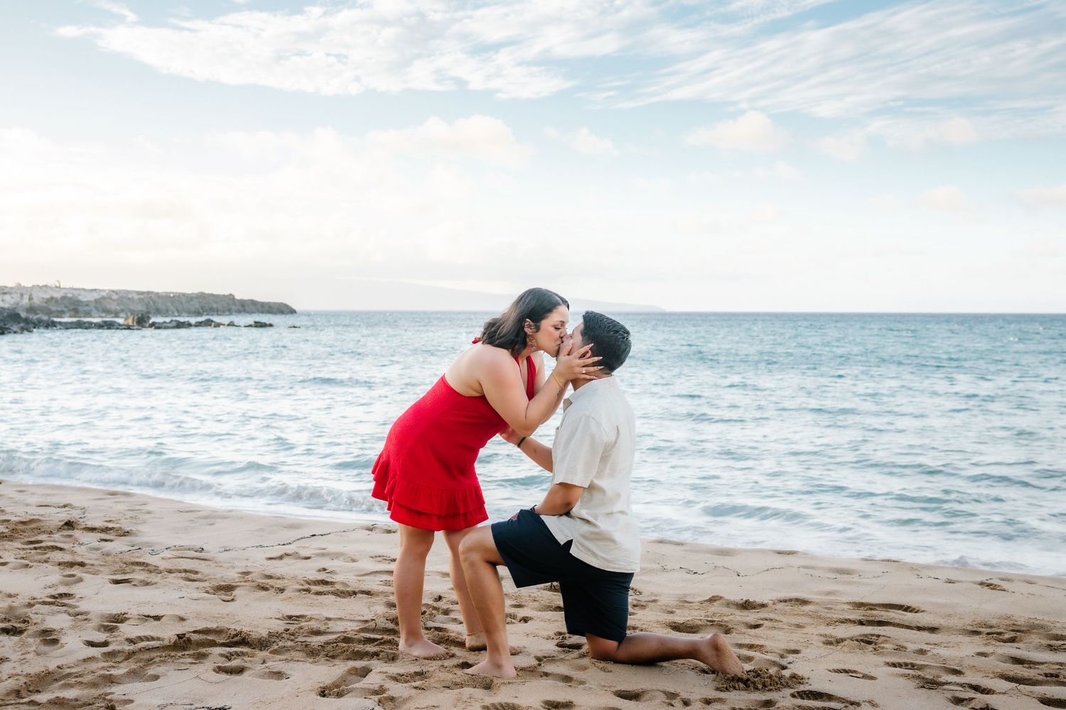 Couple sharing a romantic kiss on a sandy beach, man on one knee proposing to woman in red dress.