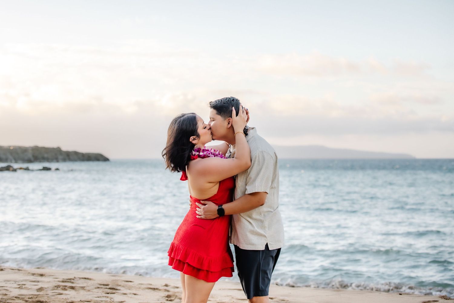 Couple kissing on a sandy beach at sunset with ocean waves and distant islands in the background.