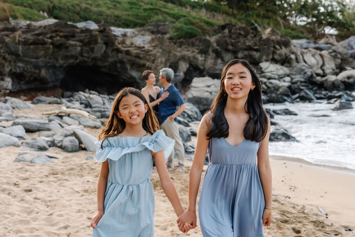 Two young girls in matching light blue dresses hold hands smiling on a beach with rocky cliffs behind them.