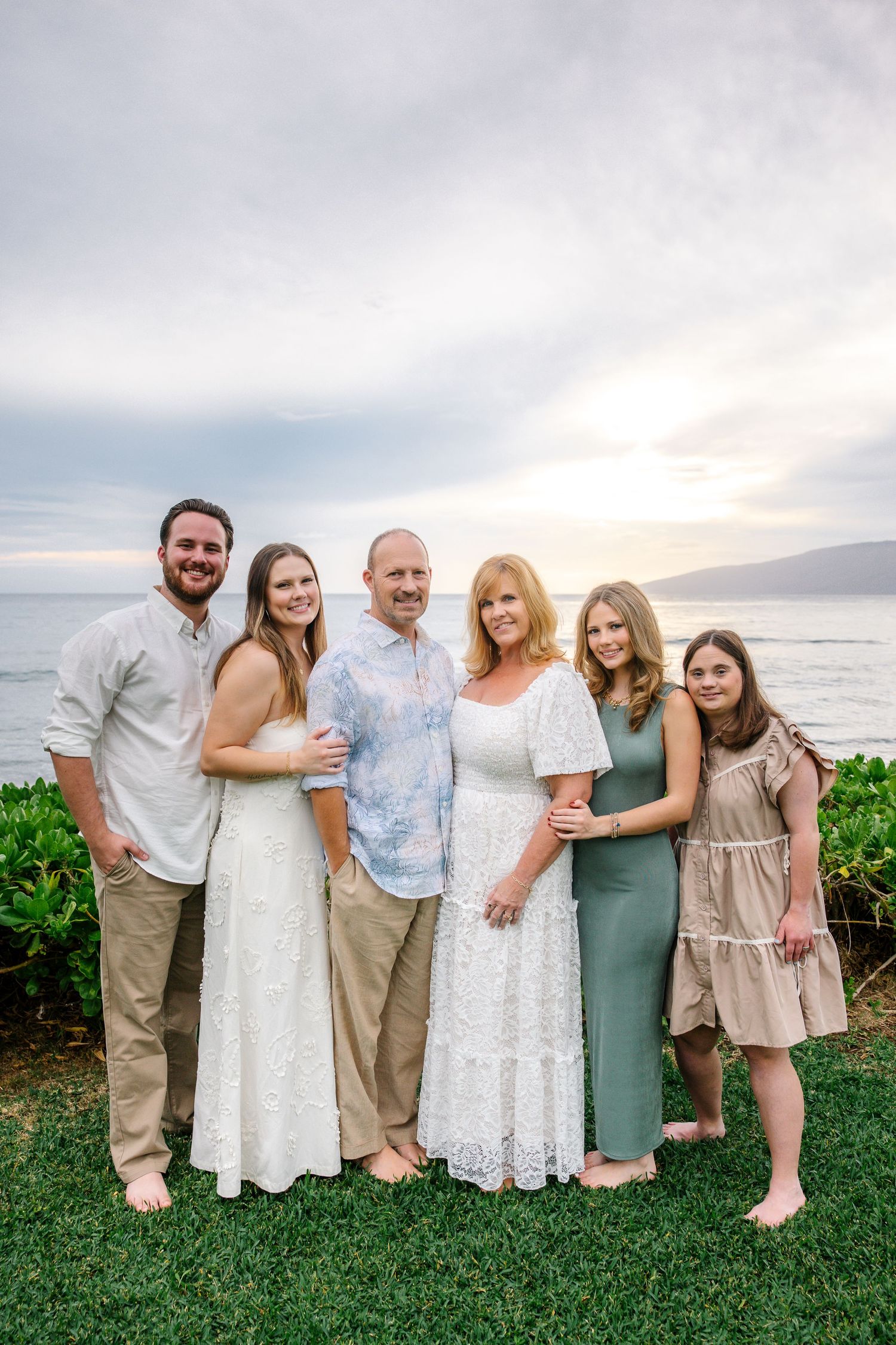 Family group poses together on a grassy oceanfront lawn in Hawaii wearing coordinated white and neutral outfits.