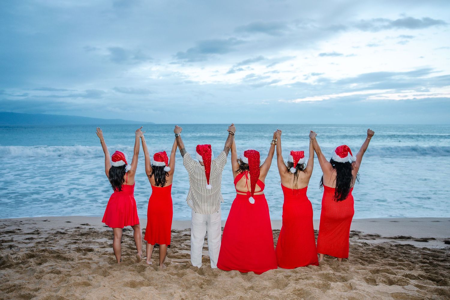 Wedding party in red dresses celebrating with raised arms on a beach at sunset.