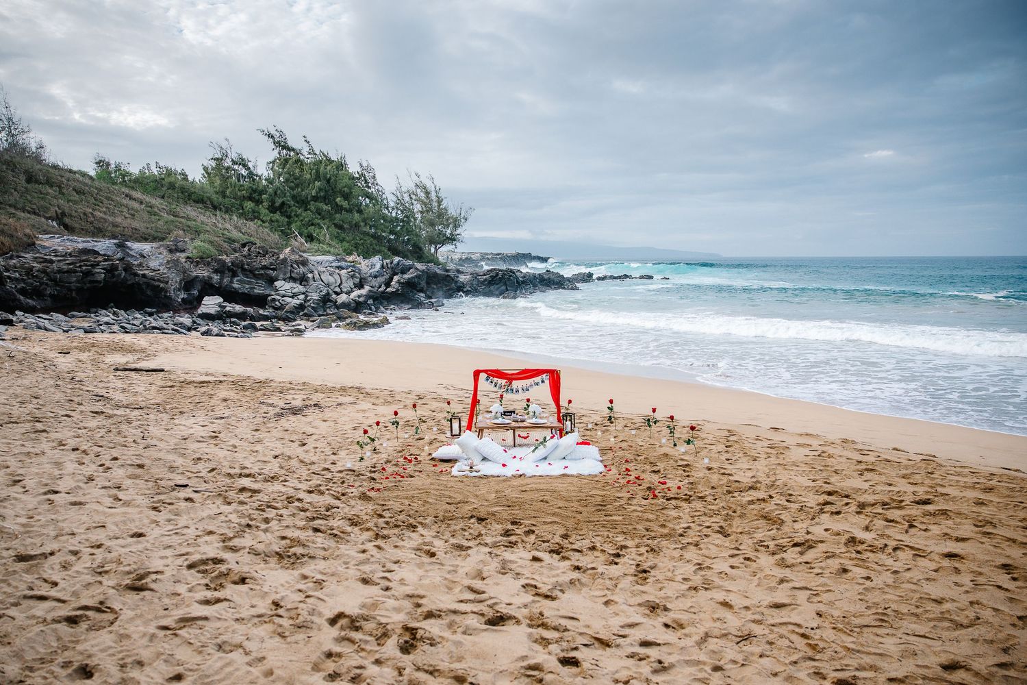 A red wedding arch decorated with flowers stands on a sandy beach with rocky cliffs and ocean waves in the background.