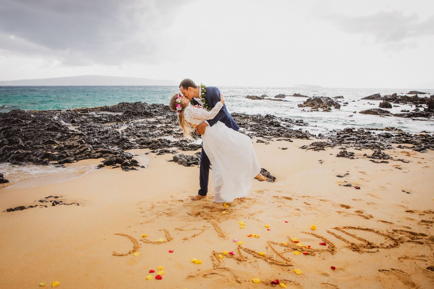 Couple sharing a romantic kiss on a sandy beach with words written in the sand during their Hawaii wedding.