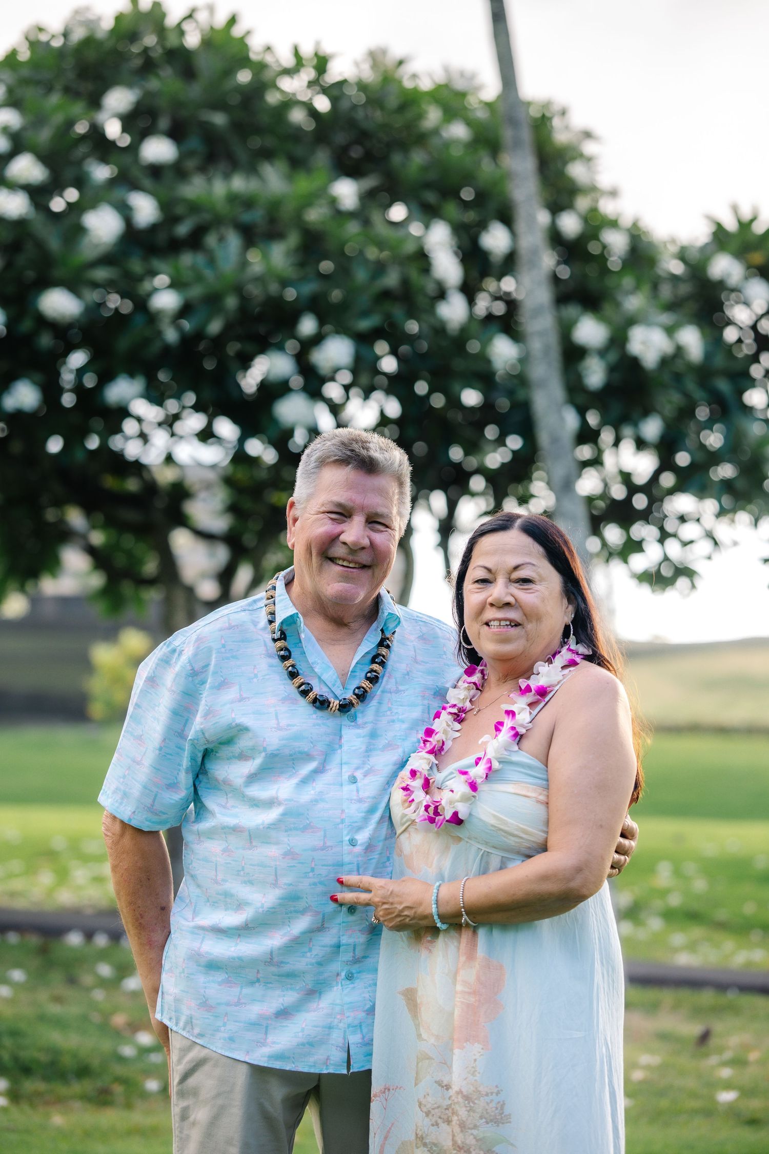 Smiling couple wearing leis poses outdoors in Hawaii with lush green trees in the background.