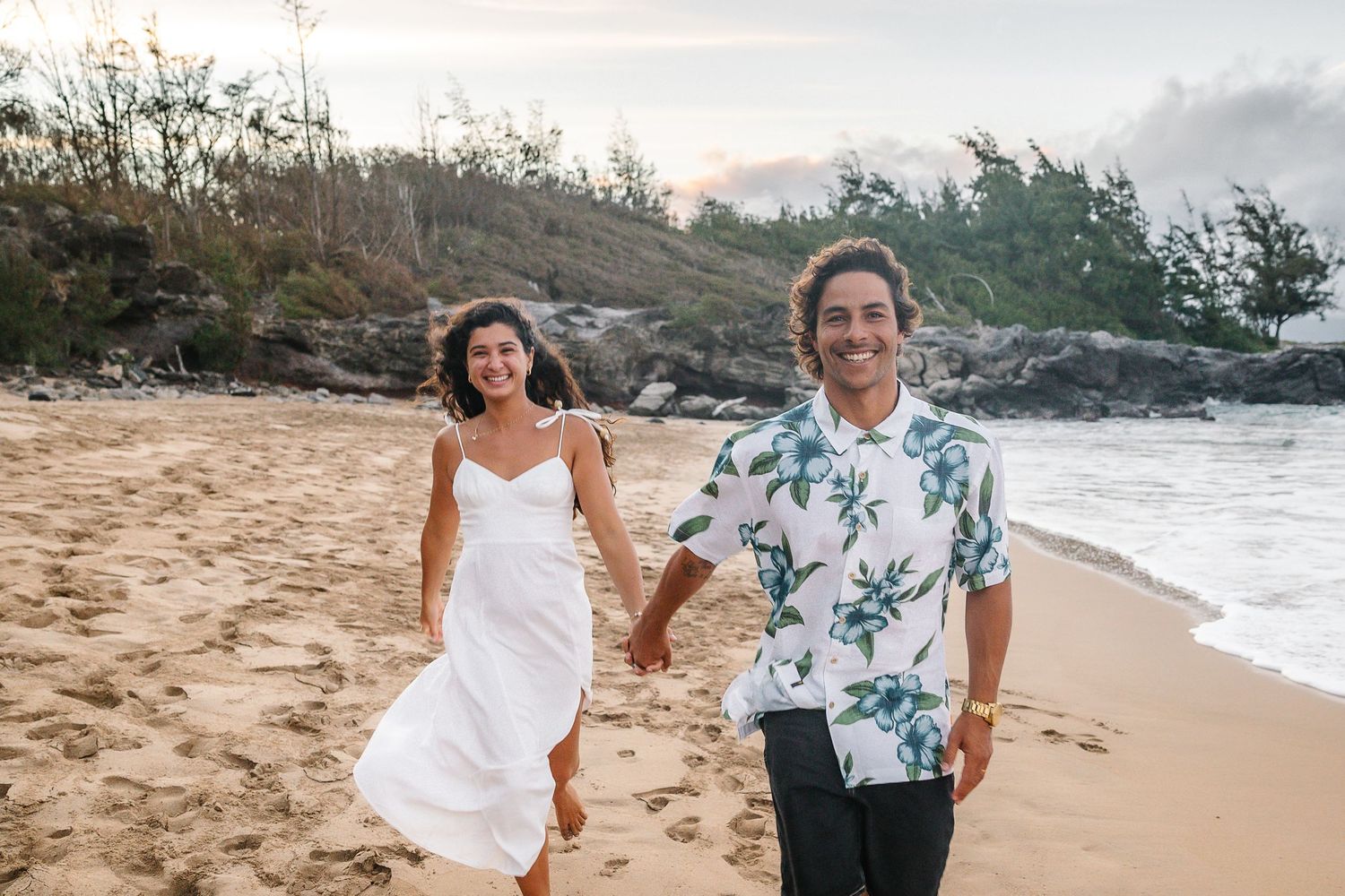 Couple walks hand in hand along a scenic beach with rocky cliffs in the background.