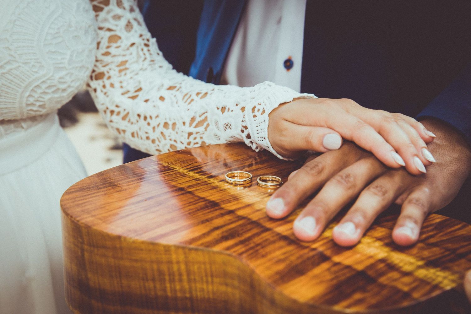 Wedding rings rest on a polished wooden ukulele surface with lace dress detail.
