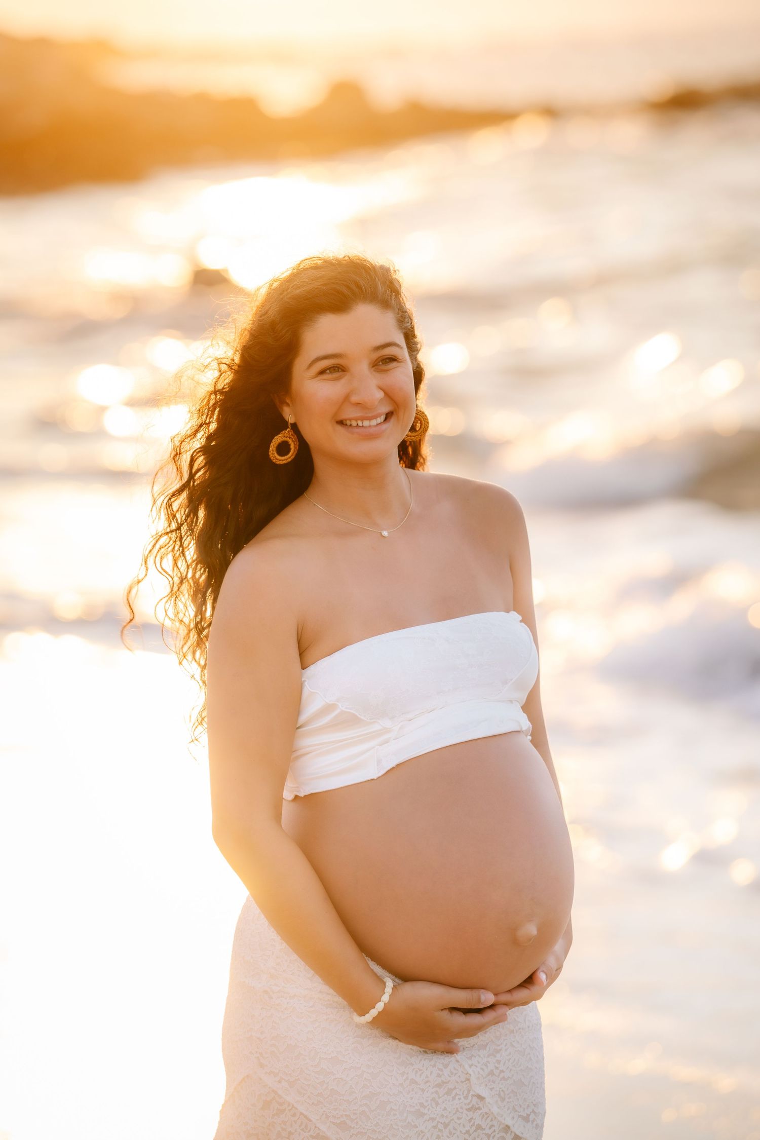 Smiling pregnant woman in white strapless top holding her belly on a beach at golden sunset.