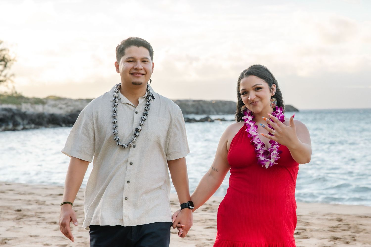 Couple holding hands on a Hawaiian beach at sunset, man in beige shirt with lei, woman in red dress showing engagement ring.