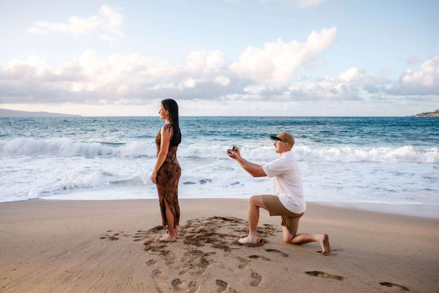 Man proposing to woman on a sandy beach with ocean waves and cloudy sky backdrop.