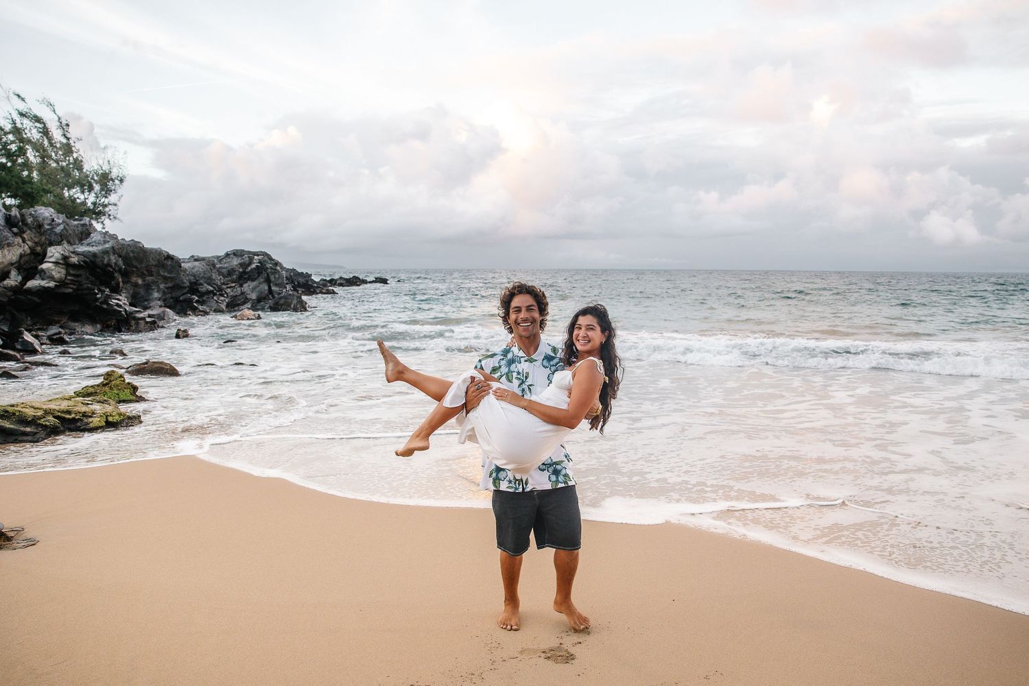 Couple plays and dances together during a beach photoshoot with waves crashing behind them.