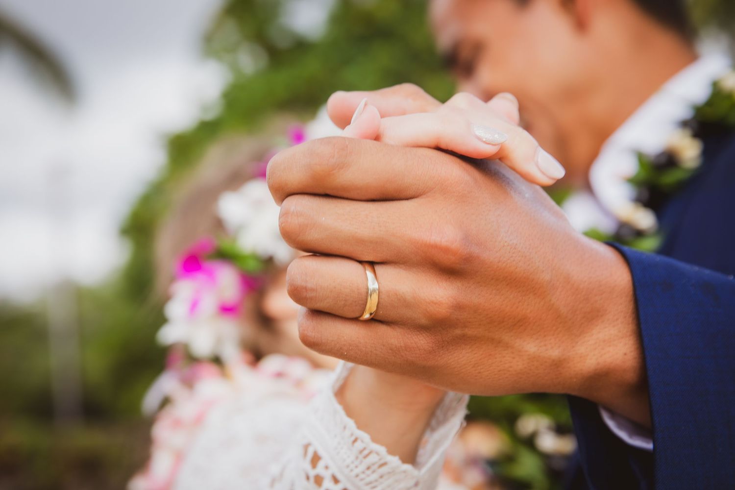Close-up of newlyweds holding hands, gold wedding band visible, with floral bouquet in background.