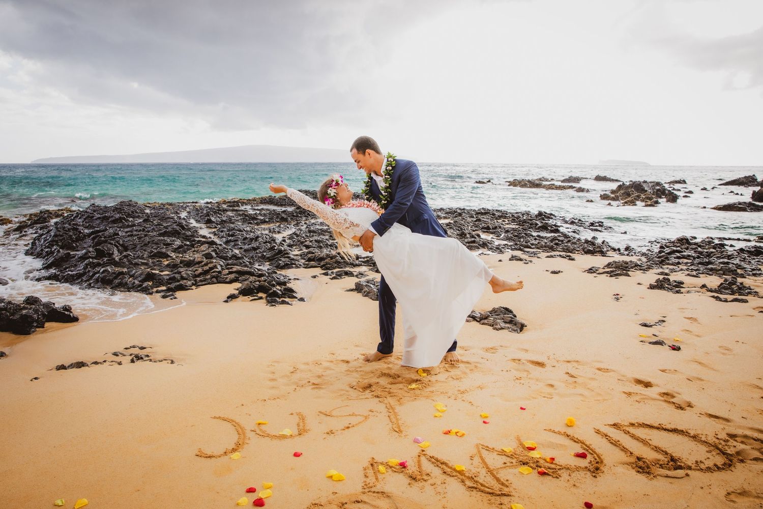 Bride and groom share romantic dip kiss on sandy Hawaii beach with ocean waves and rocks in background.