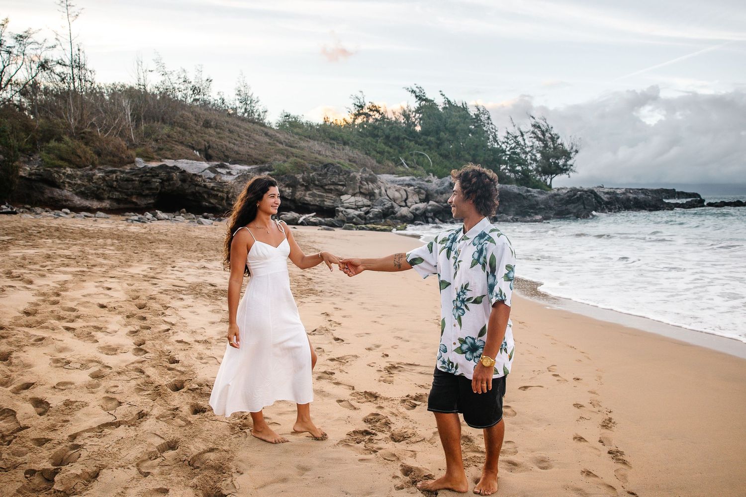 A romantic couple shares intimate moments on a scenic beach with rocky cliffs and ocean waves at sunset.