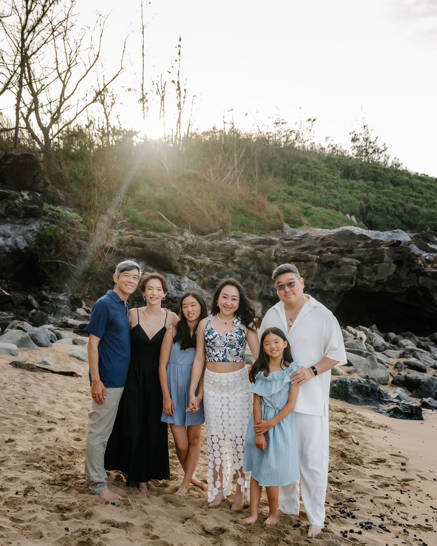 Family of six posing together on a sandy beach at golden hour with rocky cliffs and glowing sunset behind them.