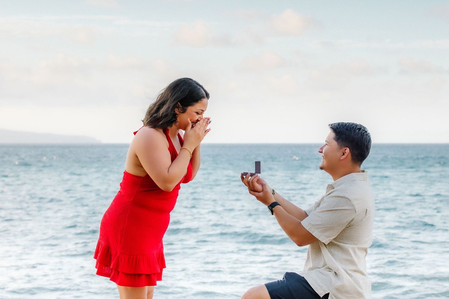 Man proposes to woman on beach, she covers mouth in surprise, holding ring box by ocean.