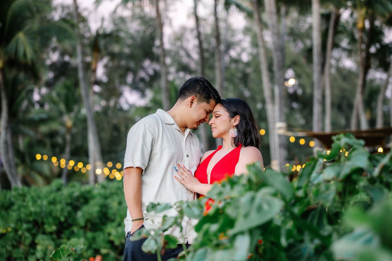 Romantic couple sharing an intimate moment in a lush tropical garden with bokeh lights and palm trees.
