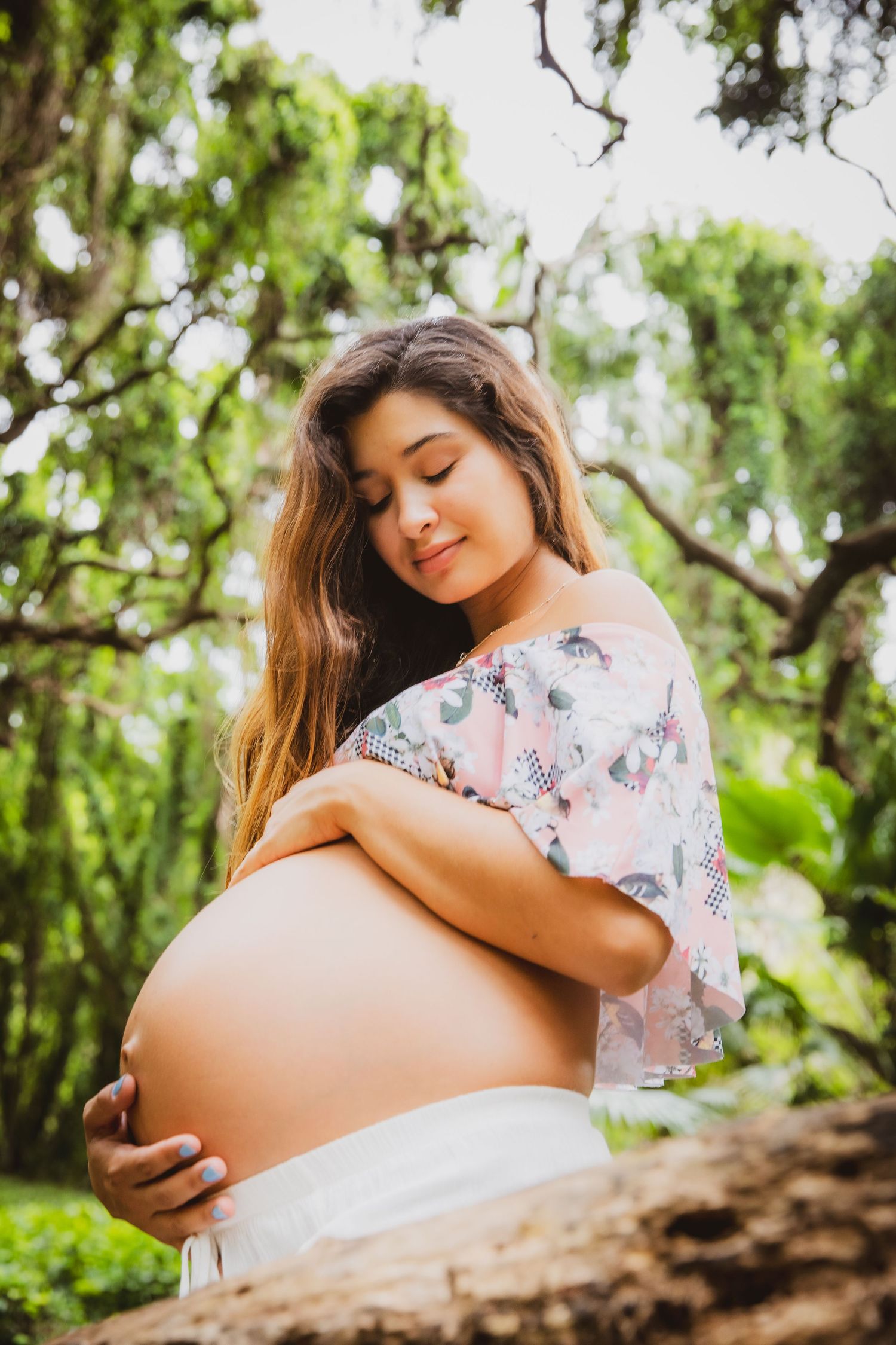 Pregnant woman in floral top cradling her bare baby bump outdoors among lush green trees.