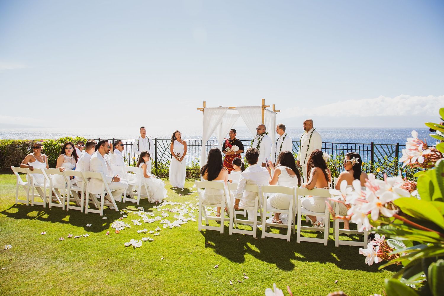 Outdoor wedding ceremony on a lush green lawn with white chairs, bamboo arch, and ocean views under a clear blue sky.