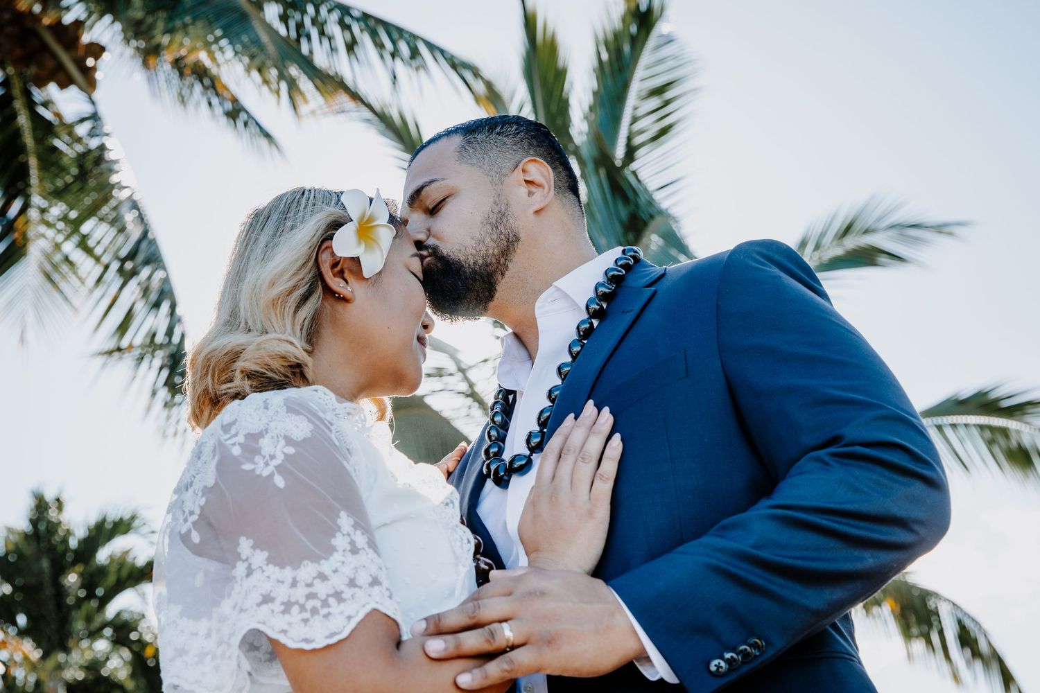 Couple sharing a romantic kiss at tropical beach wedding, bride wearing floral hair accessory, groom in blue suit.