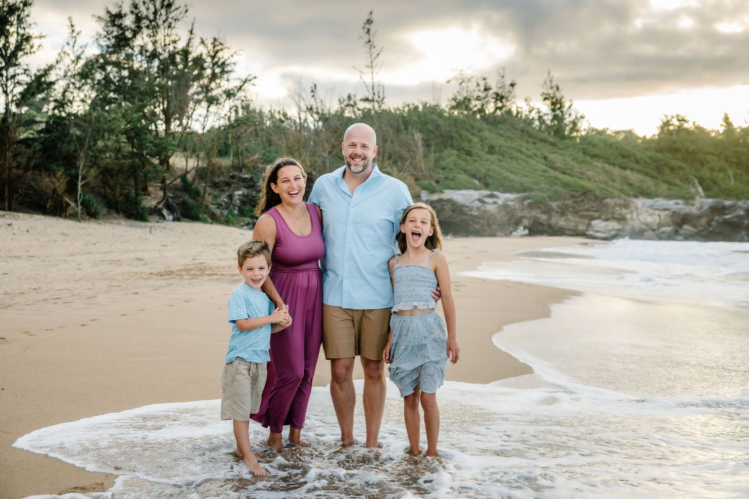 Family of four smiling on a sandy beach at sunset, waves at their feet, lush greenery in the background.