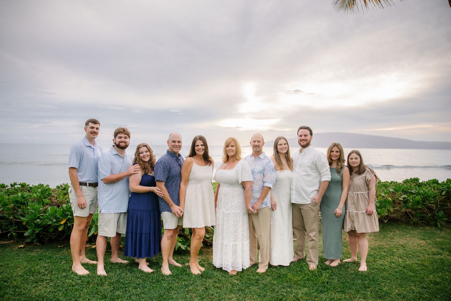 Large family group portrait on a grassy oceanfront setting in coordinated neutral attire.