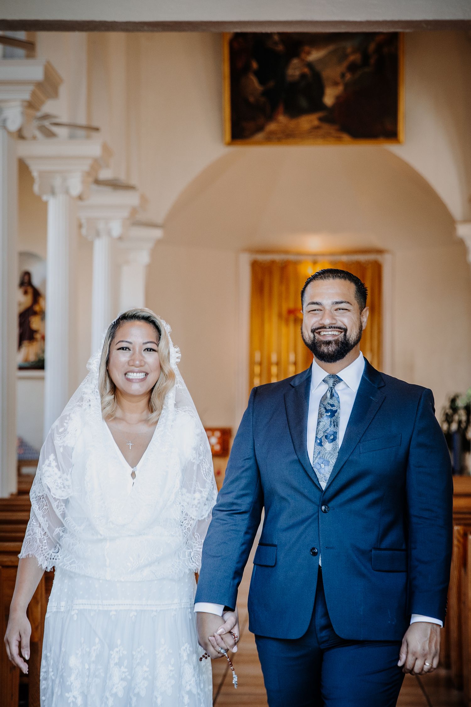 Smiling bride in lace gown and veil holds hands with groom in navy suit inside a church aisle.