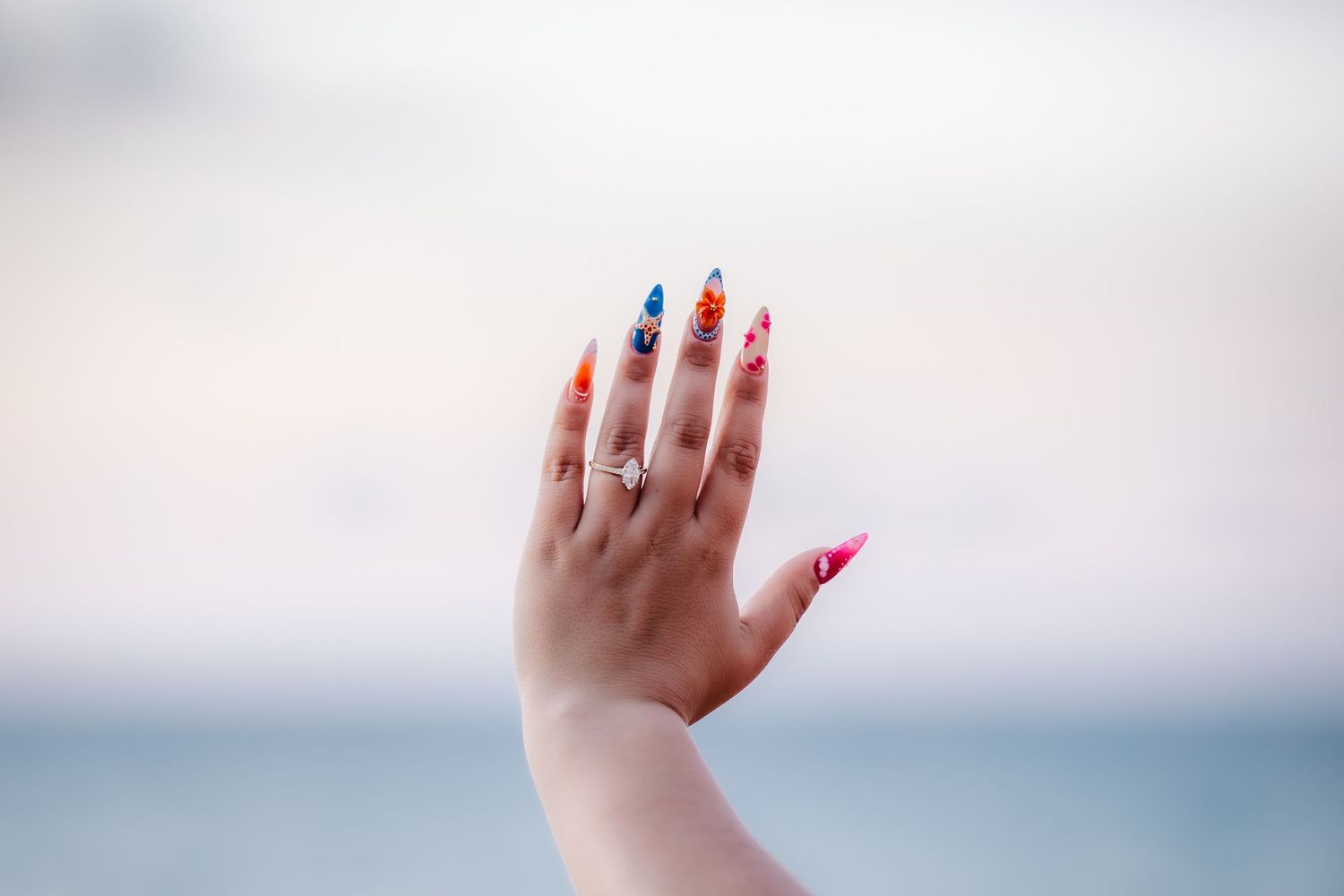 Hand with long stiletto nails featuring colorful floral nail art designs and a delicate ring, set against a blurred ocean backdrop.