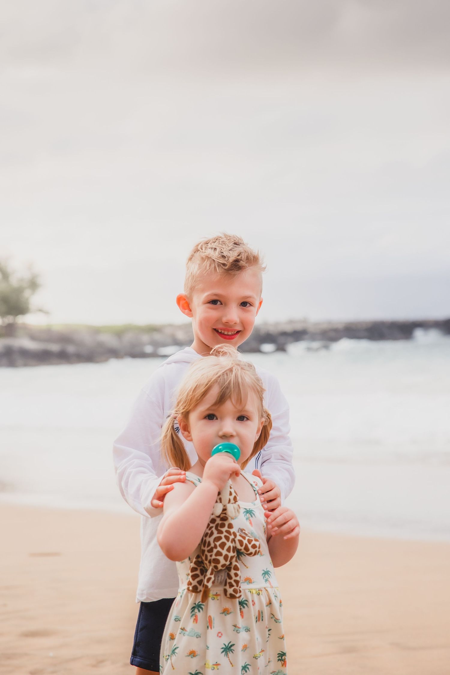 Two young children pose on a sandy beach, boy in white hoodie hugging girl with pacifier holding giraffe toy.