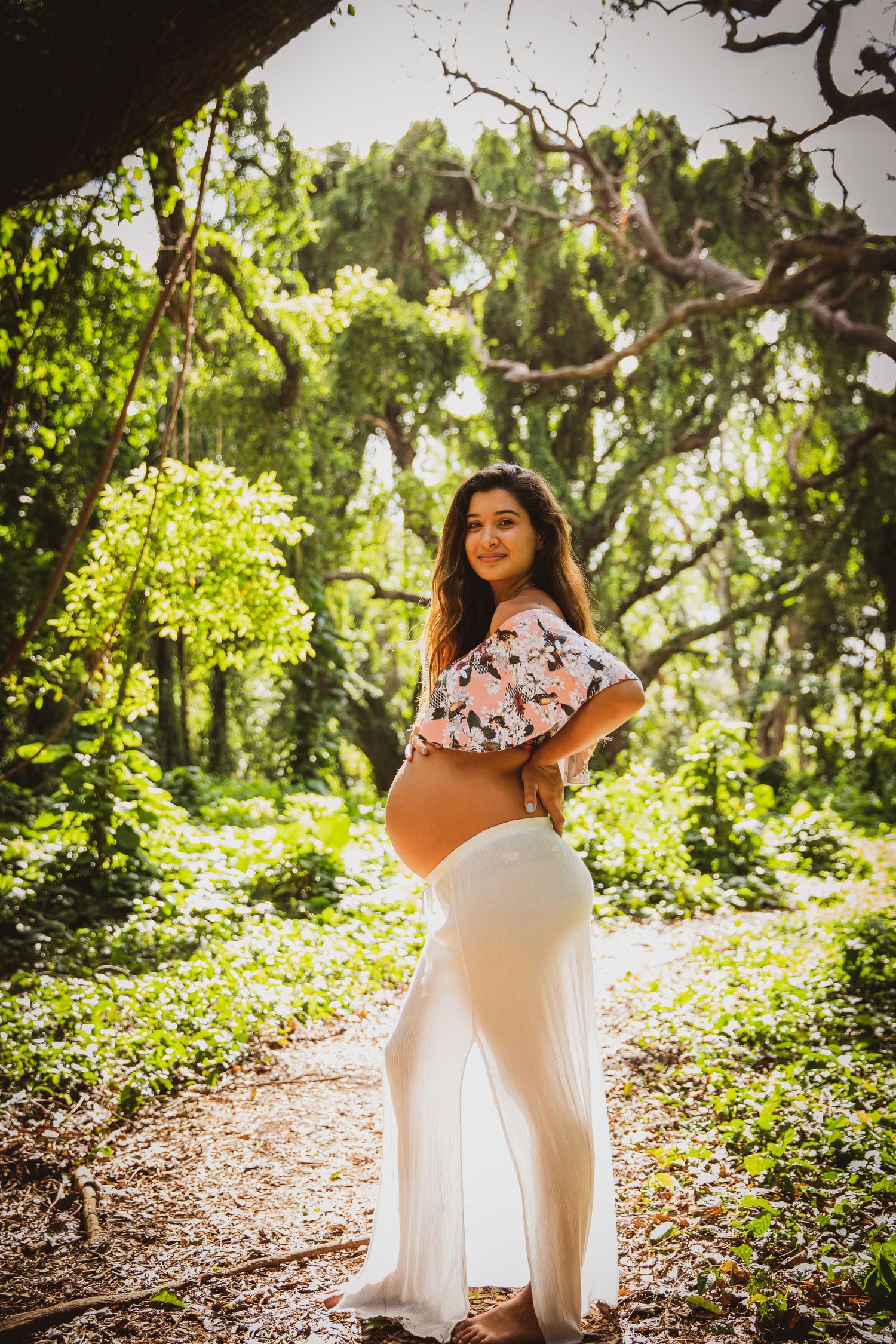 A maternity photoshoot in a lush green forest setting with natural sunlight filtering through the trees.