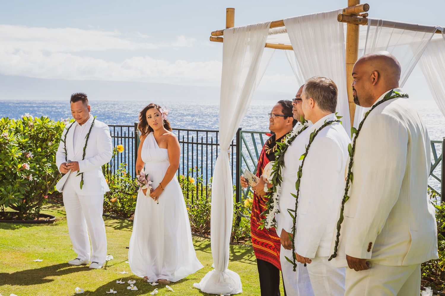 Outdoor Hawaiian wedding ceremony with bride and groom at bamboo arch, ocean view backdrop, guests in white attire.