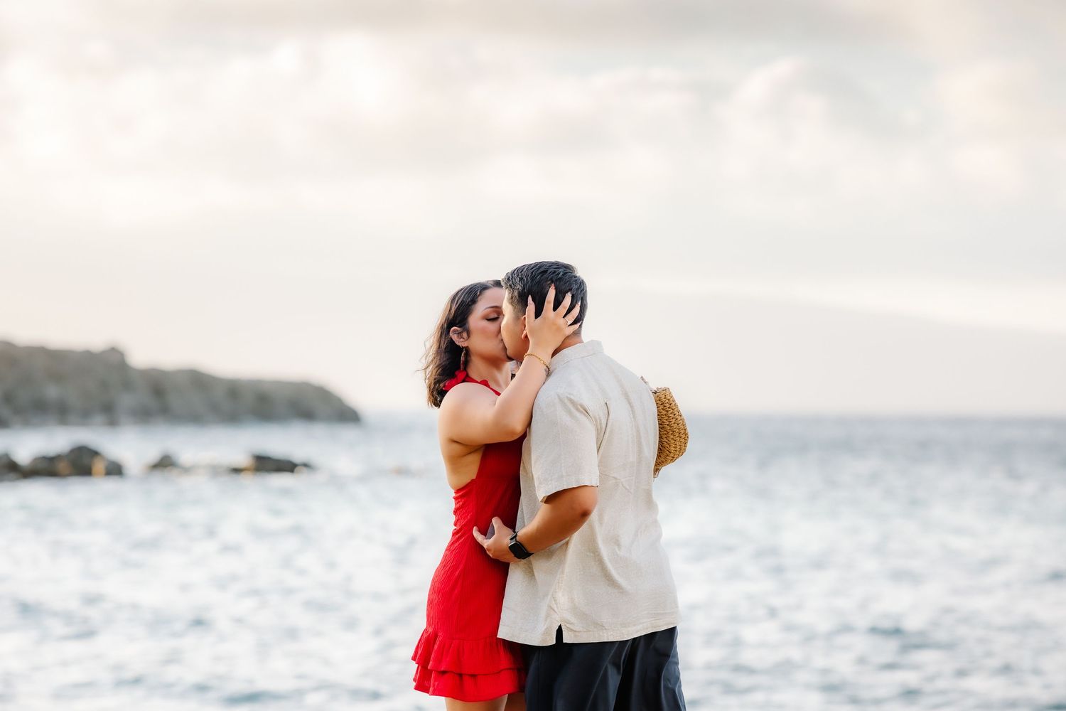 Couple sharing a romantic kiss on a rocky beach with ocean waves and cloudy sky in the background.