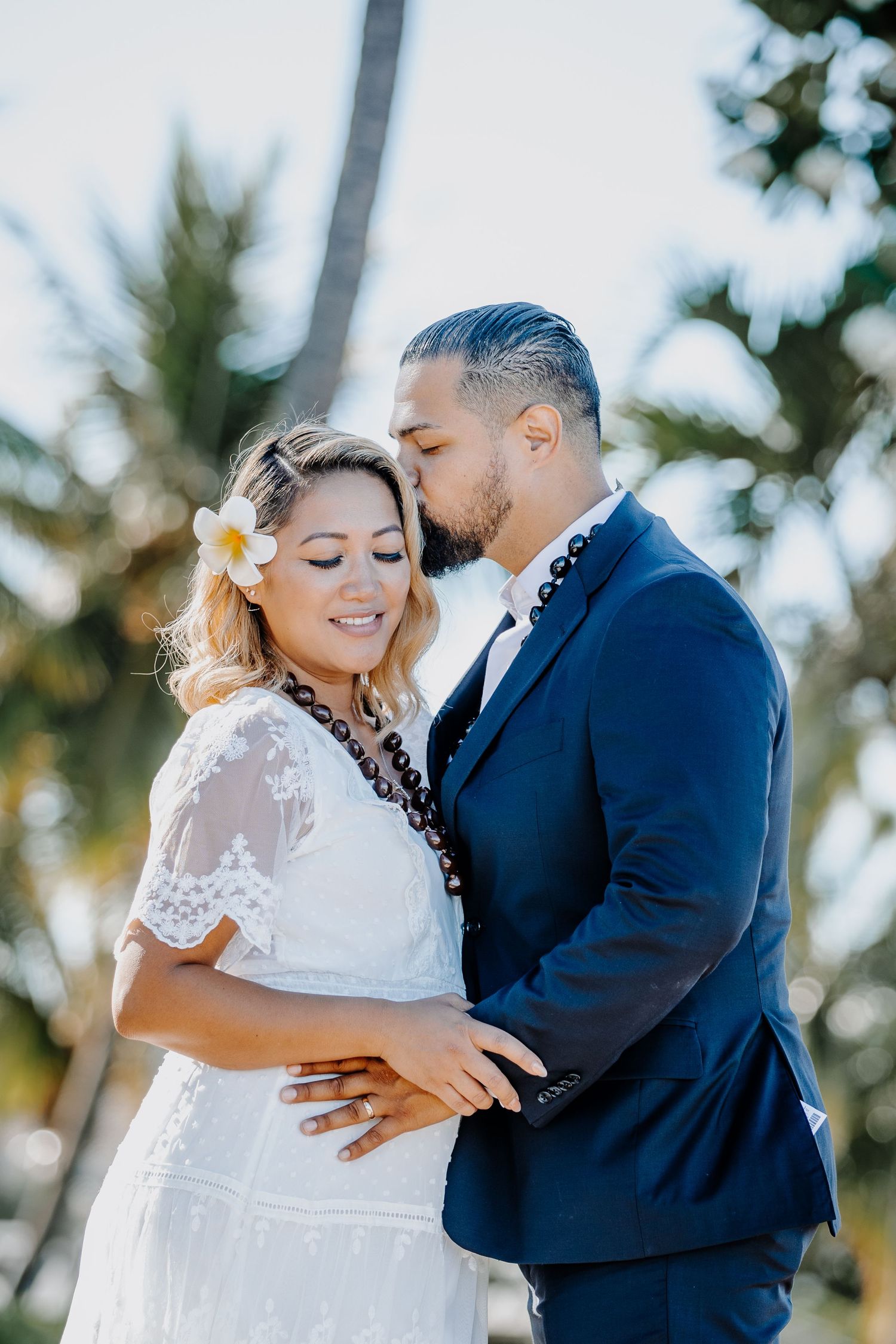 Couple in wedding attire sharing a romantic kiss outdoors, with palm trees in the background on a sunny day.