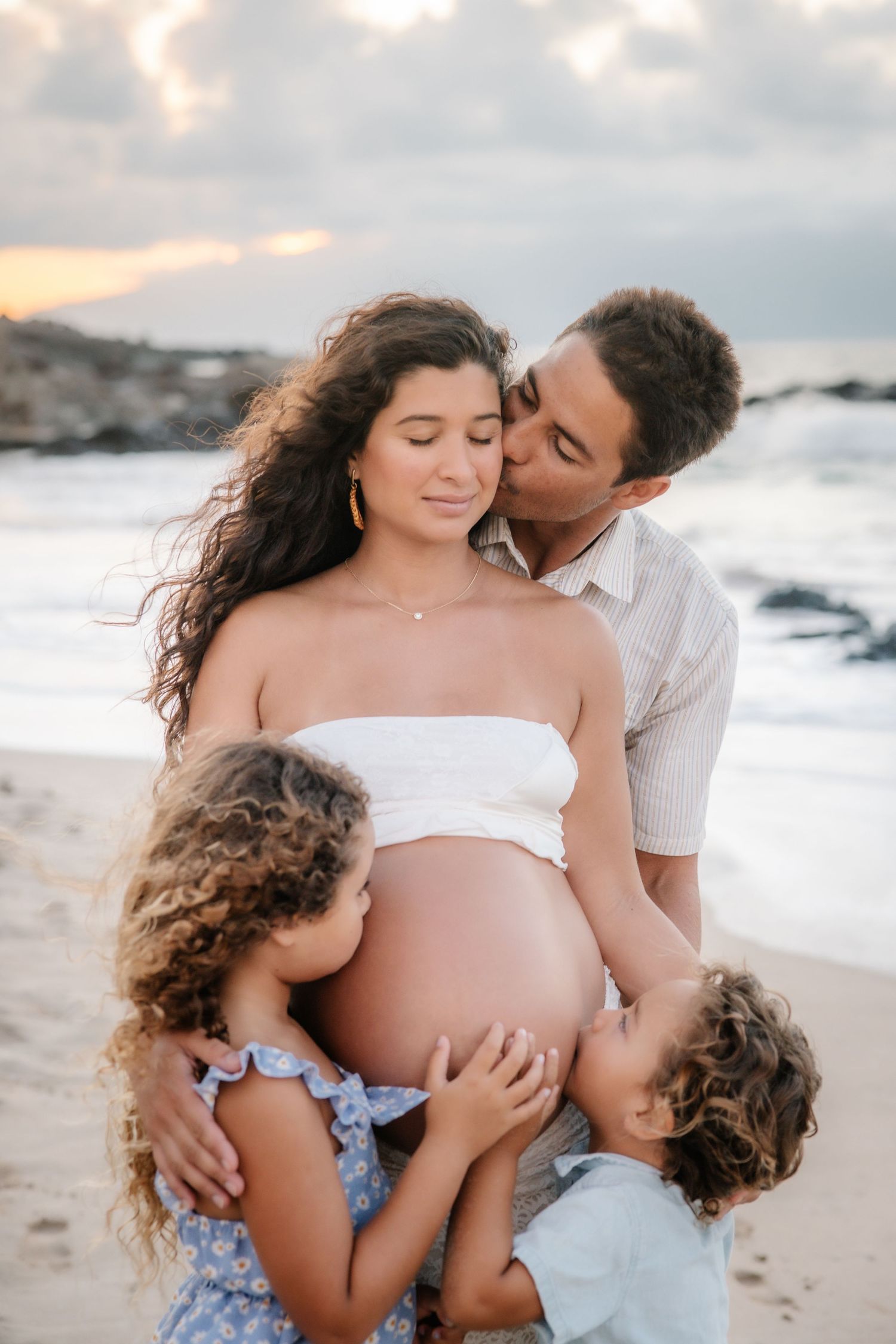 Pregnant woman on beach with husband kissing her cheek and two children hugging her baby bump at sunset.