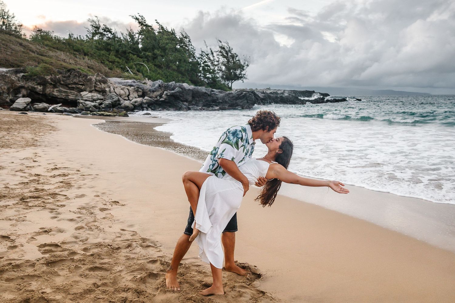 Playful beach engagement photos with dramatic coastal landscape and crashing waves.