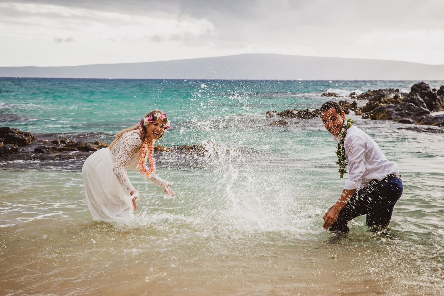 Playful newlyweds splashing water at each other on a tropical beach.