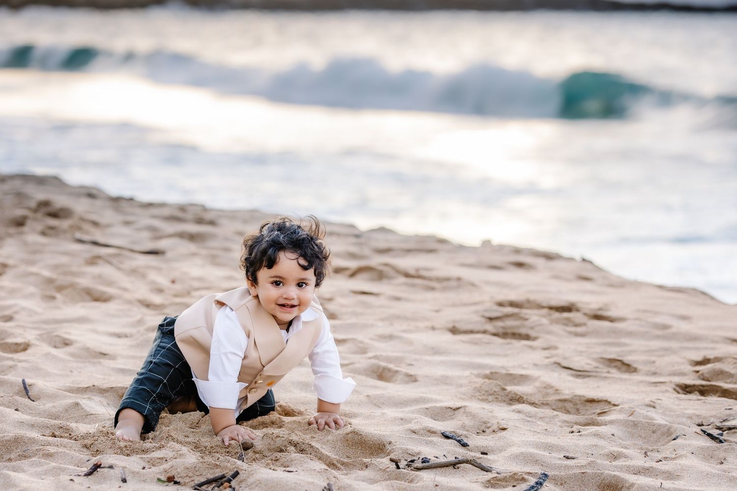 A happy toddler in a white shirt crawls on sandy beach with ocean waves in the background.