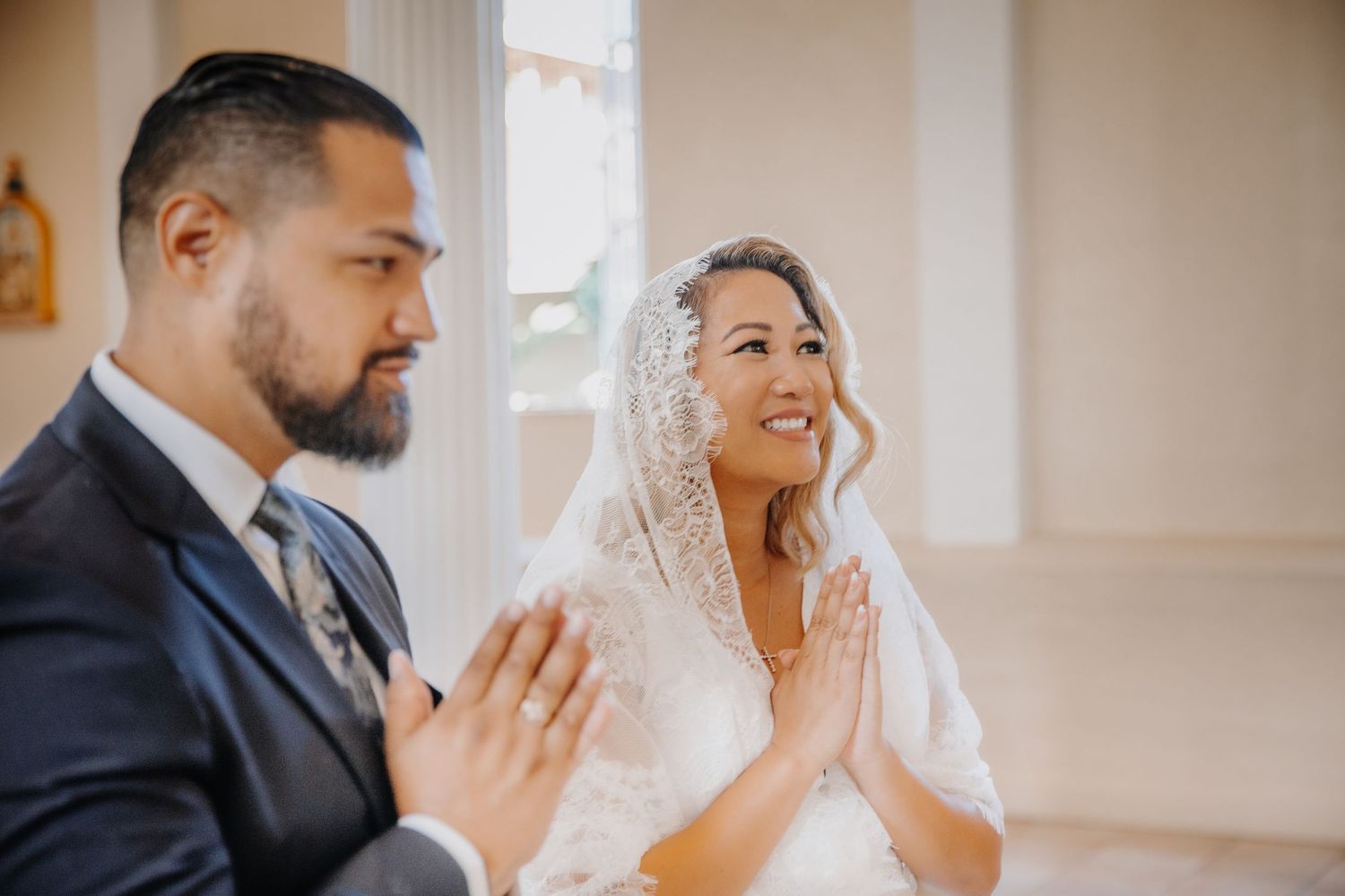 Bride and groom praying together at wedding ceremony, bride wearing lace veil, groom in navy suit.