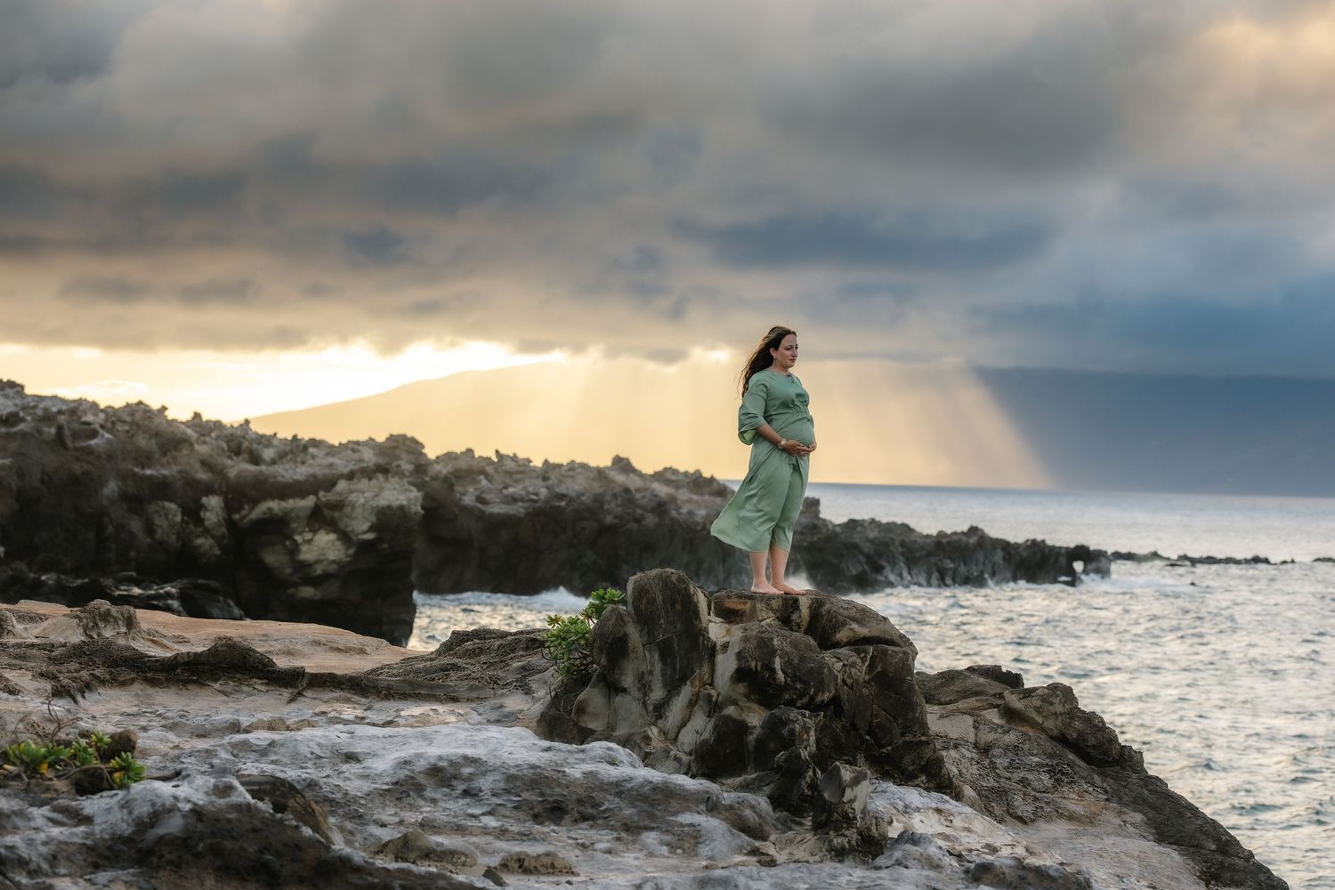 Pregnant woman in green dress stands on rocky shoreline with dramatic stormy sky and sunbeams over ocean.