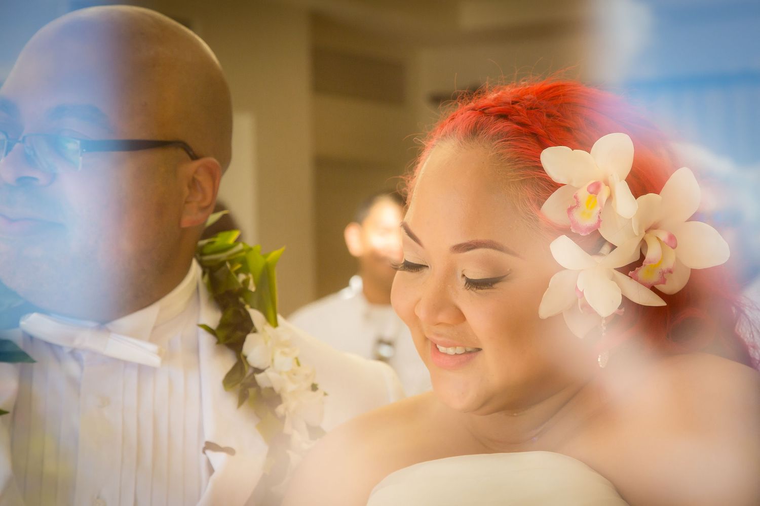 Bride with pink hair adorned with white orchids smiles beside groom wearing green lei at tropical wedding ceremony.