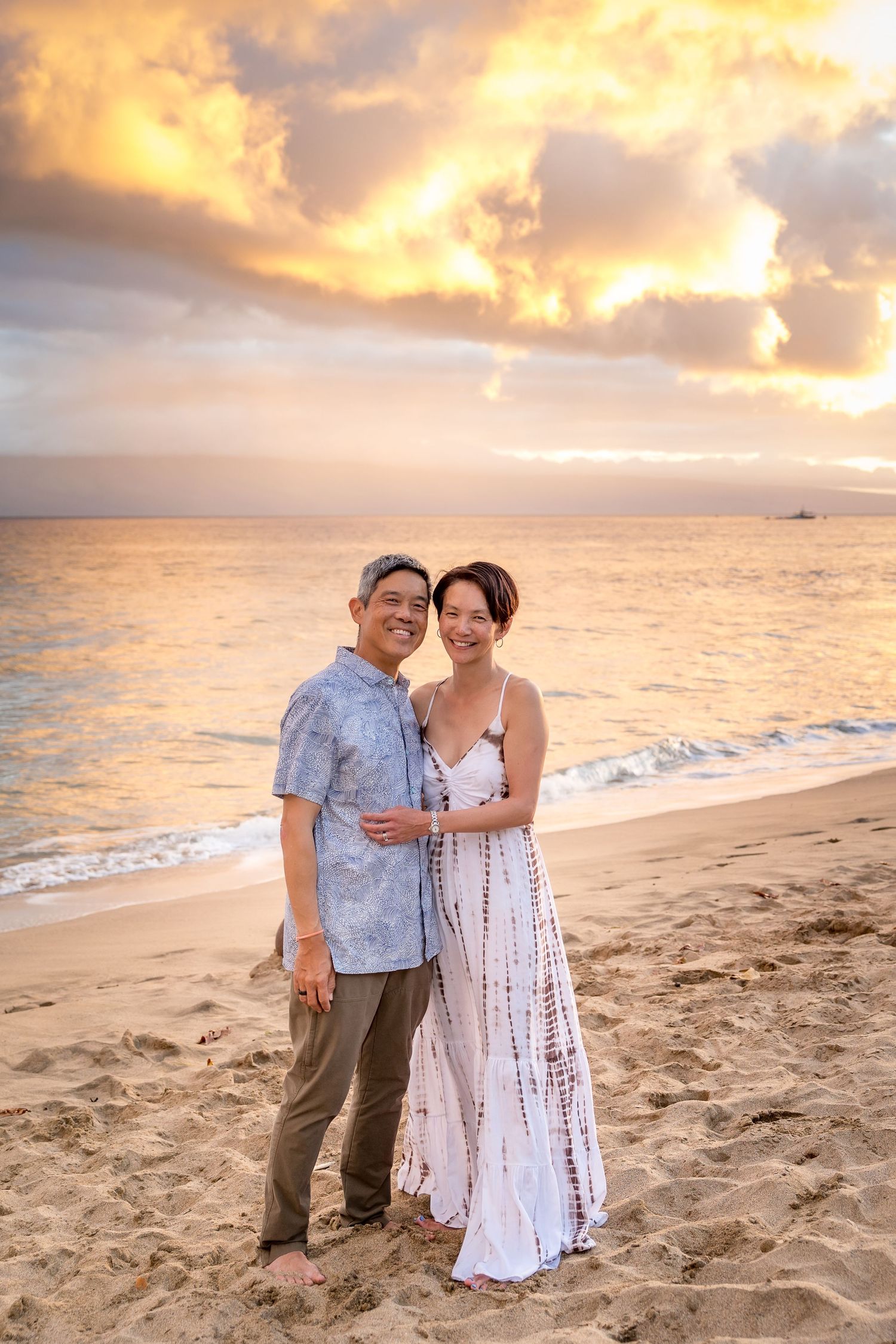 A couple stands together on a sandy beach with a dramatic orange and pink sunset sky.