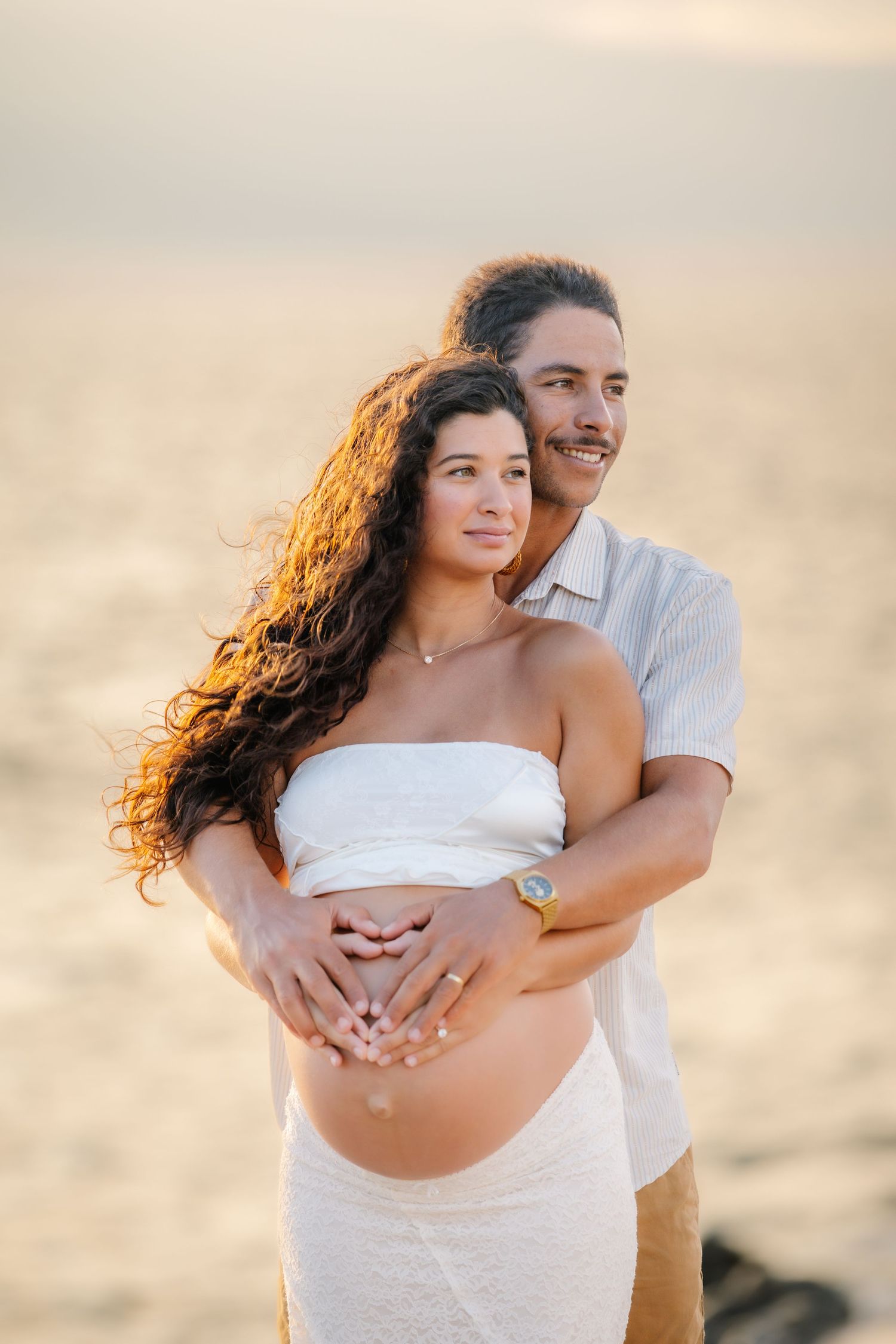 Couple embracing on beach at sunset, woman with curly hair in white tube top showing pregnant belly, man hugging from behind.