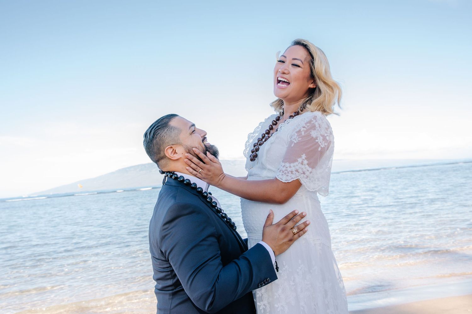 Couple laughing and embracing on a beach in Hawaii, woman in white lace dress, man in navy suit.