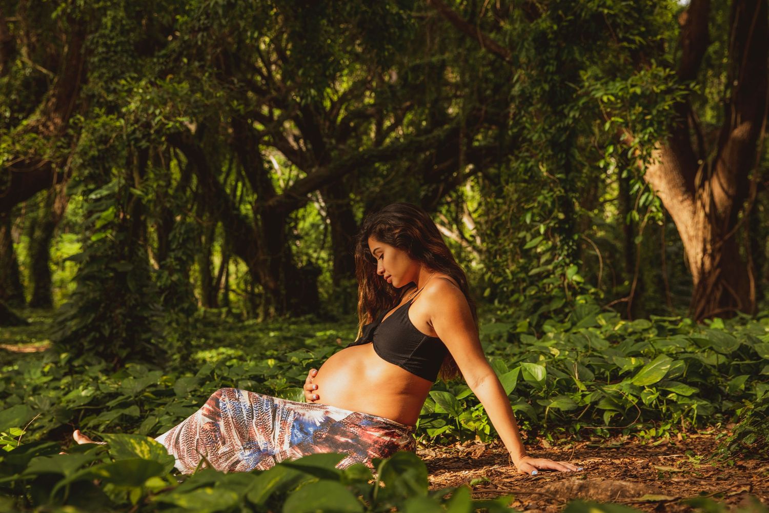 Pregnant woman resting on forest floor surrounded by green foliage.