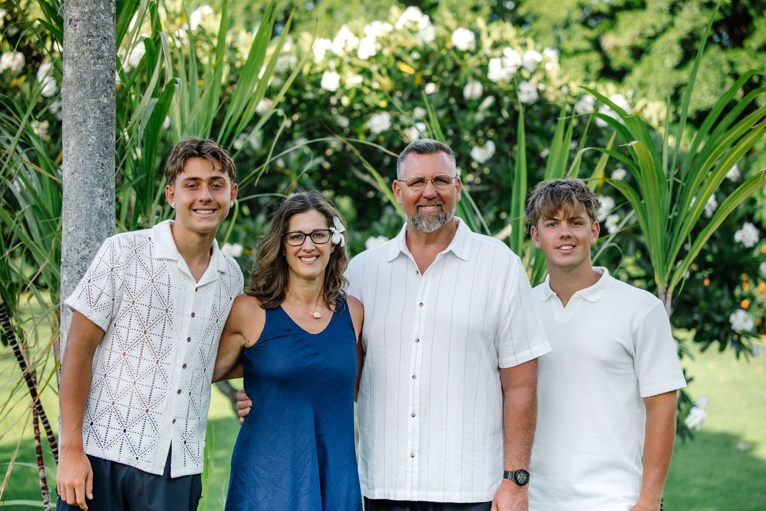 Family of four smiling outdoors among tropical palm trees and white flowers on a sunny day.