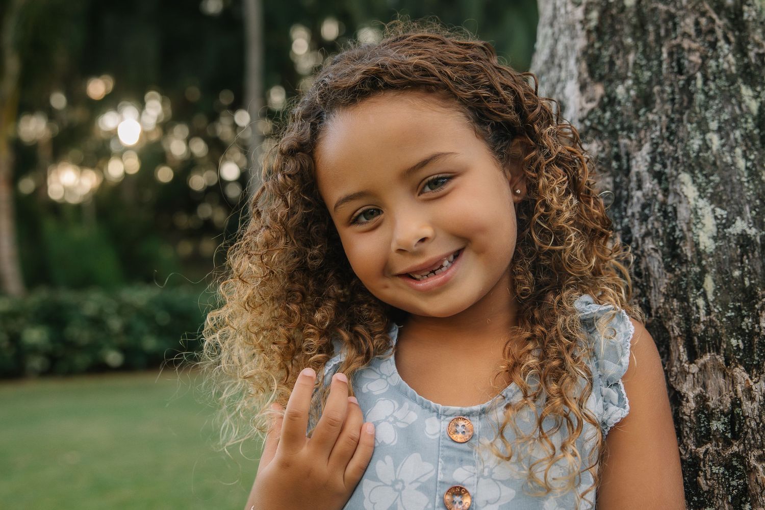 A young child with curly hair smiles while wearing a blue floral dress and standing next to a tree outdoors.